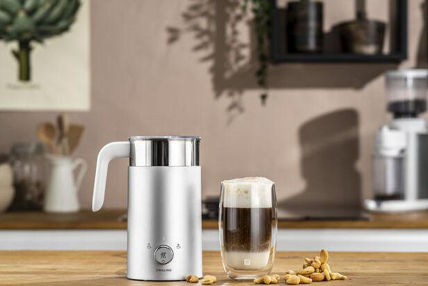 Ein weißer Milchaufschäumer und ein Glas geschichteten Kaffees mit Nüssen auf einem Holztisch. Küchenhintergrund.