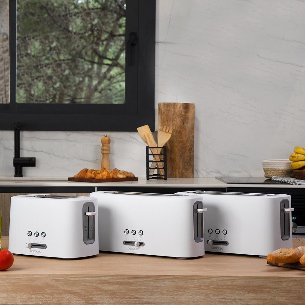 Tres tostadoras blancas sobre una mesa de madera. El fondo incluye una encimera de cocina con pan, utensilios y una ventana.