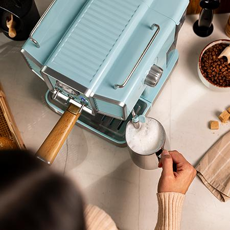 Una persona espuma leche con una máquina de espresso azul. Granos de café, cubos de azúcar y una toalla están sobre la mesa.