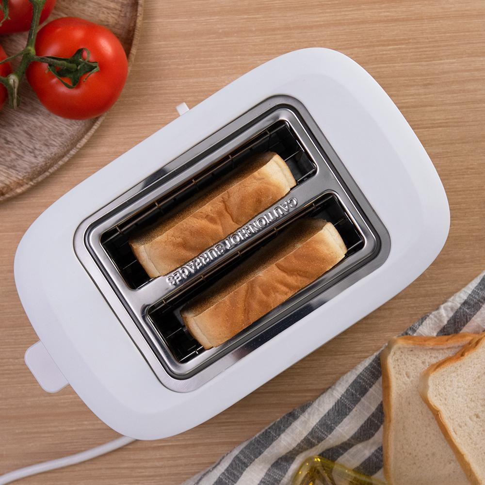 Una tostadora blanca con dos rebanadas de pan tostado. Tomates y un paño a rayas están cerca de la tostadora sobre una mesa de madera.