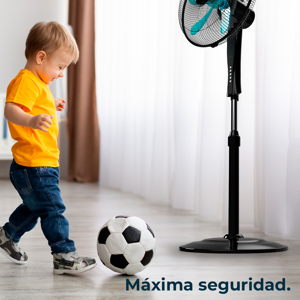 Un niño pequeño con una camisa naranja y jeans juega con un balón de fútbol cerca de un ventilador negro. La habitación tiene cortinas blancas.