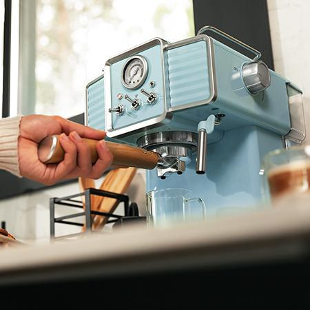 Una persona usando una máquina de espresso azul. Una mano sostiene un mango de madera. Hay vasos de vidrio y una rejilla de metal.