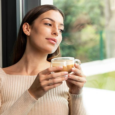 Mujer sosteniendo una taza de café, mirando por una ventana. Lleva un suéter beige. La ventana muestra follaje verde.