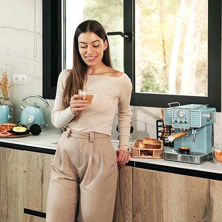 Mujer en la cocina sosteniendo un vaso. Se ven una cafetera azul, pan y una tetera. Ventana con árboles al fondo.