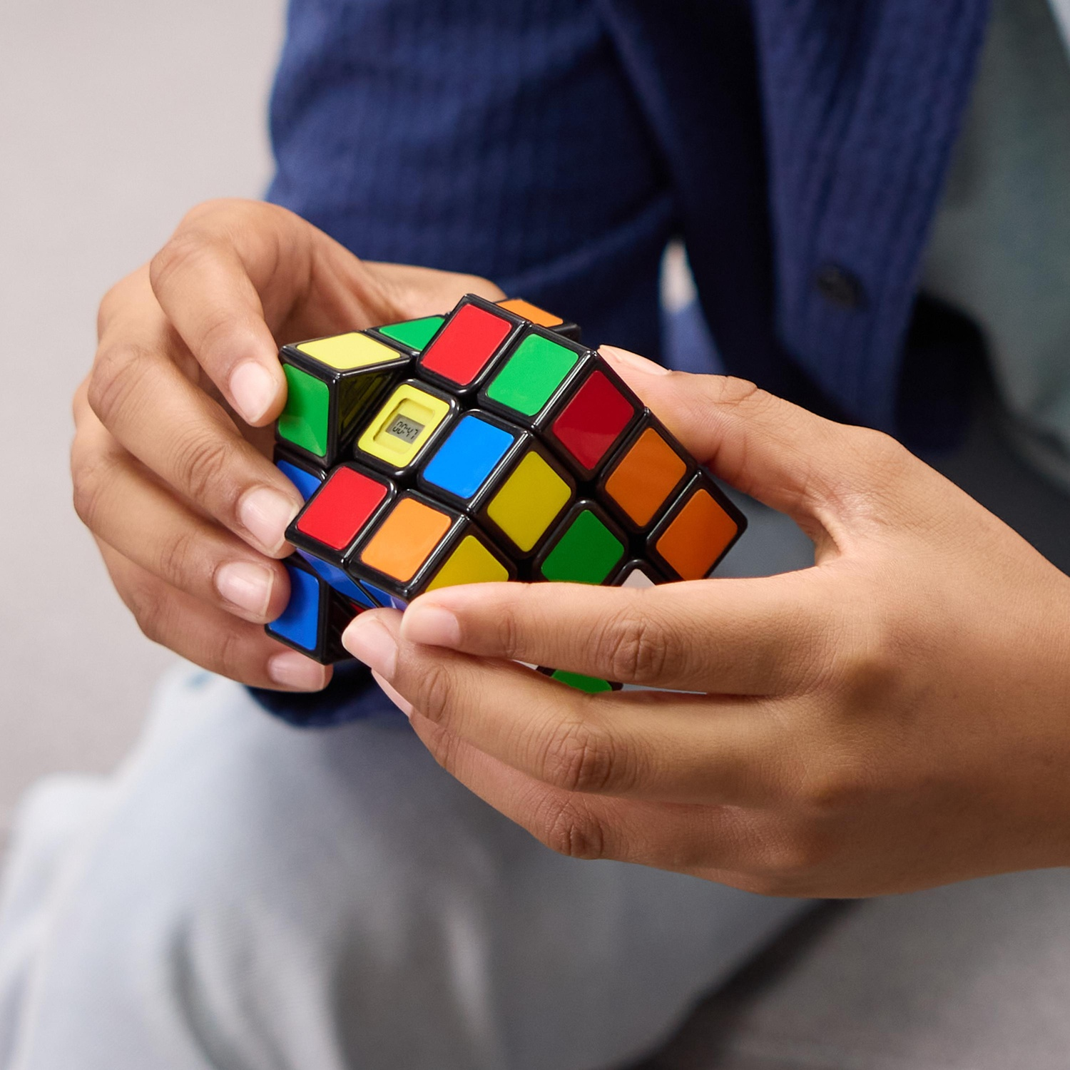 Mani che tengono un cubo di Rubik con quadrati colorati. Il cubo ha un timer digitale. La persona indossa una giacca blu.