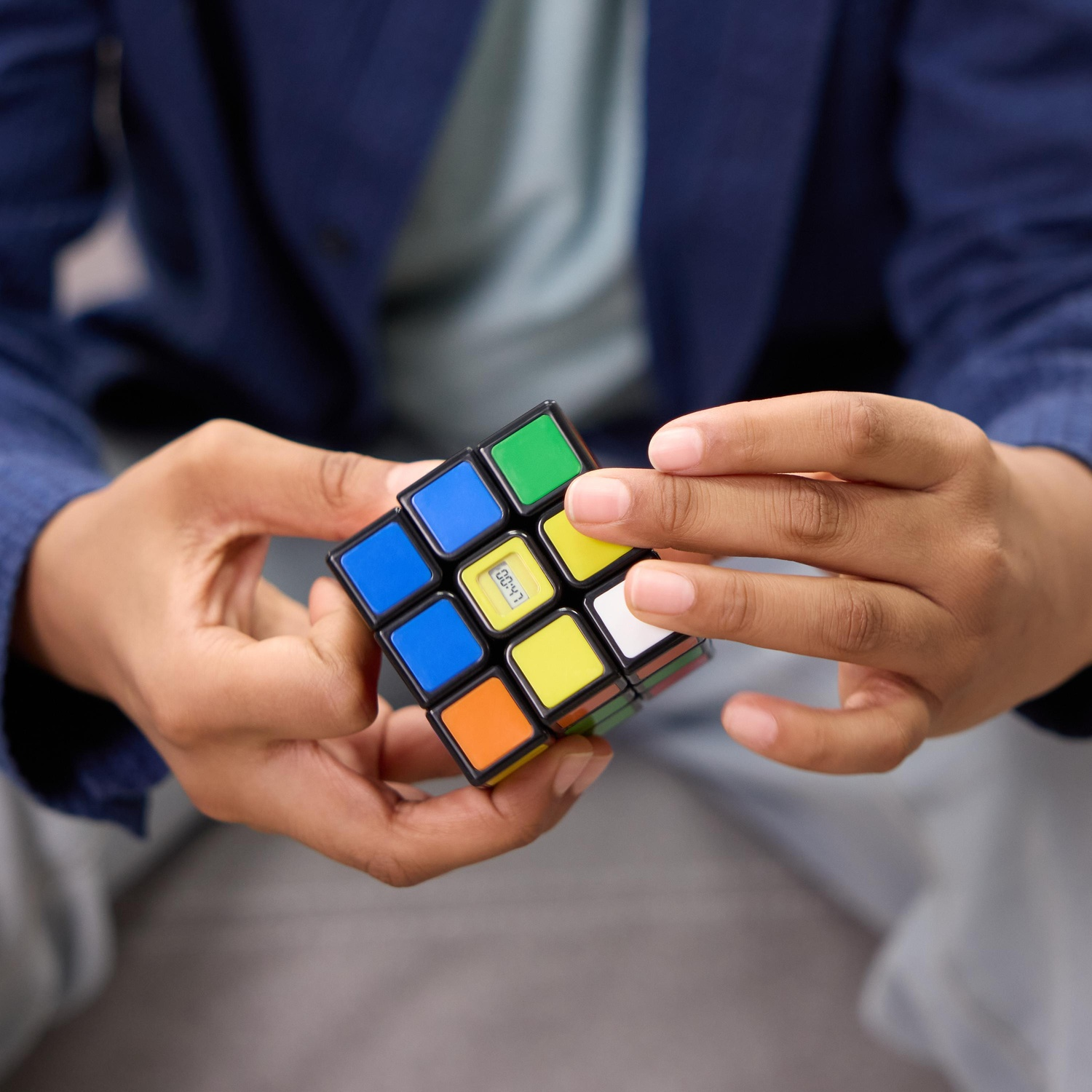 Una persona tiene in mano un cubo di Rubik con colori misti. Il cubo è a fuoco. La persona indossa una giacca blu scuro. Pantaloni grigi visibili.