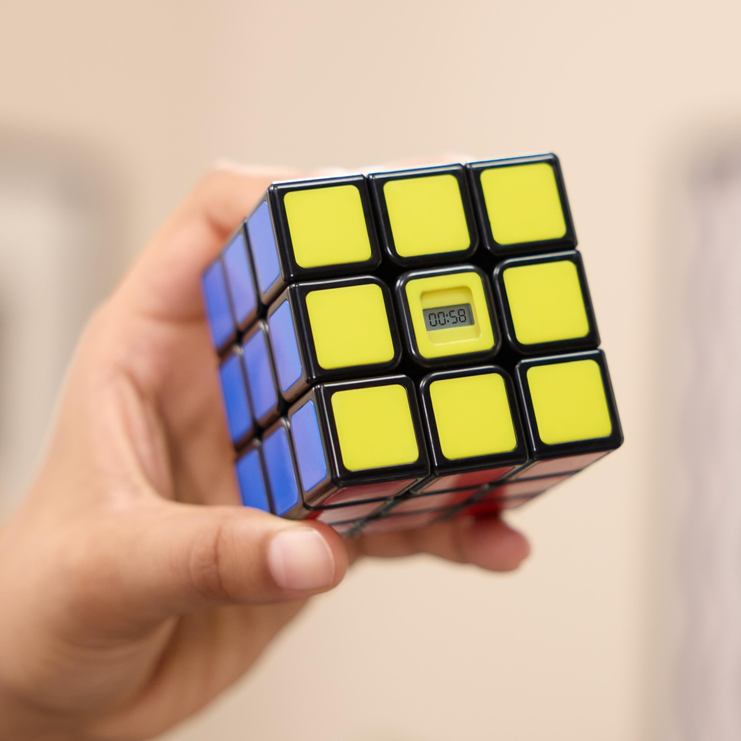 Una mano tiene un cubo di Rubik. Sono visibili lati gialli, blu e rossi. Il quadrato centrale mostra 00:58. Sfondo sfocato.