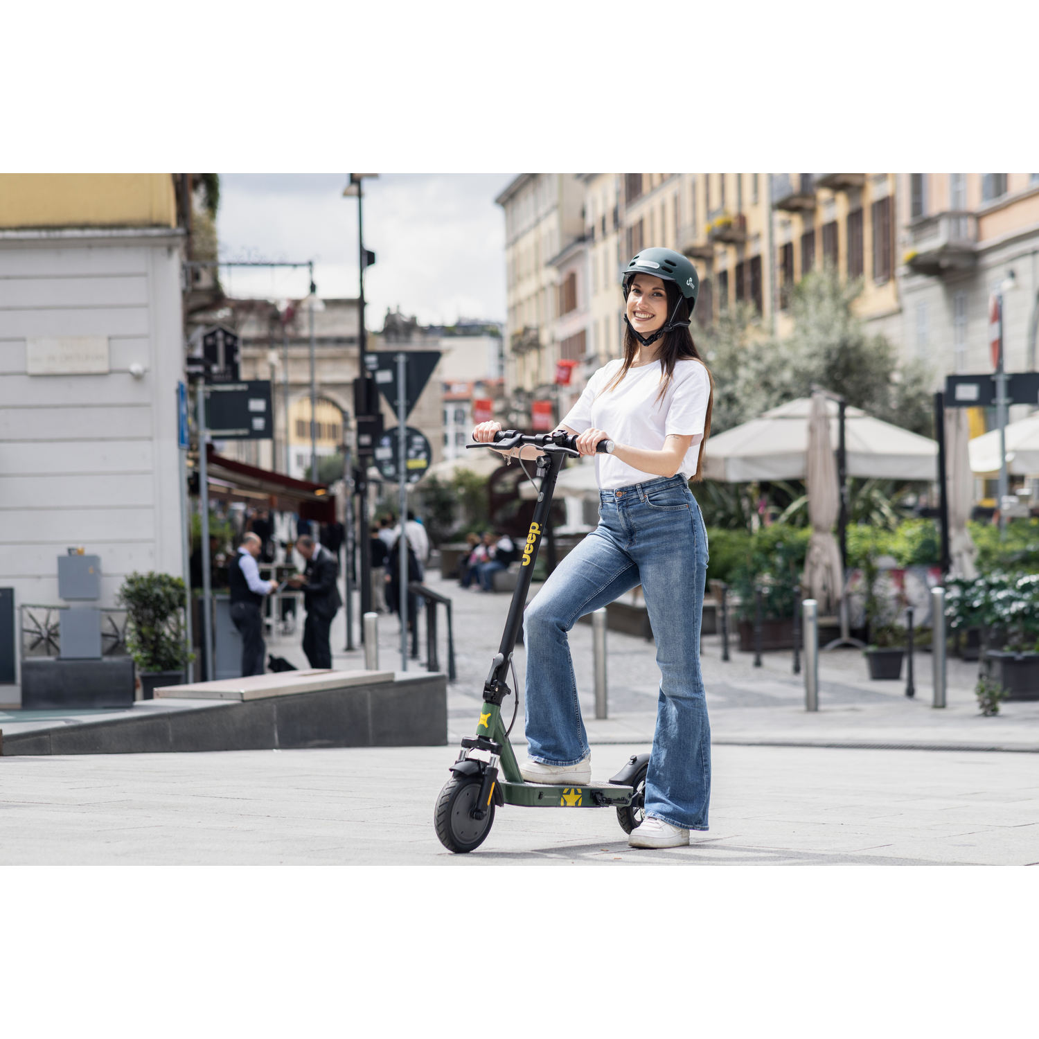 Een vrouw met helm staat op een scooter. Ze draagt een jeans en een wit t-shirt. De achtergrond is een stadsstraat met gebouwen.