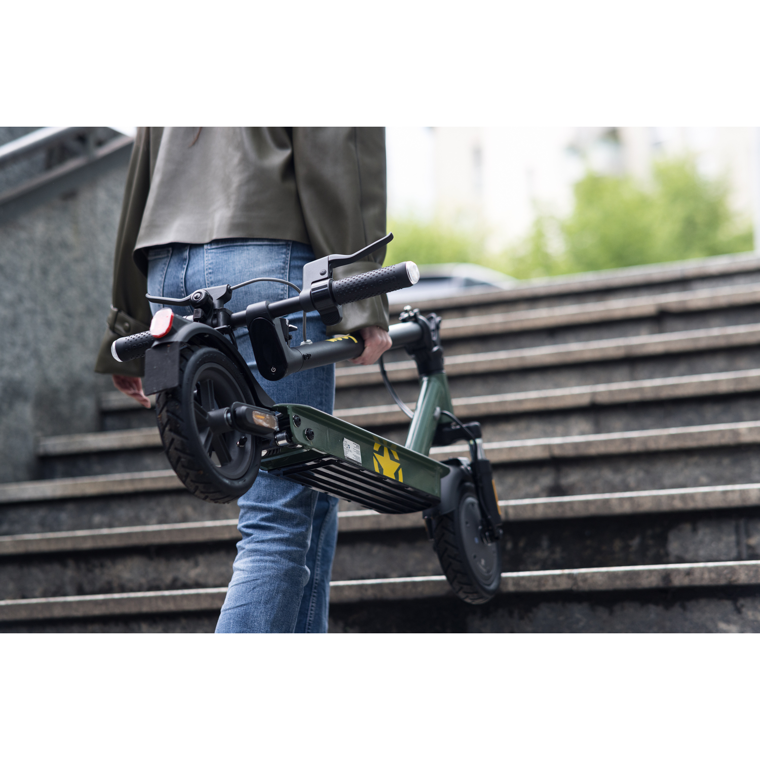 Een persoon in een spijkerbroek en jas draagt een opgevouwen groene elektrische scooter de buitentrap af. De achtergrond toont gebouwen en groen.