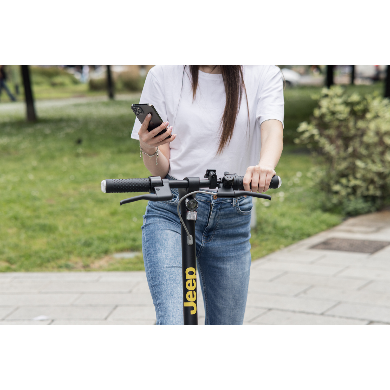 Een persoon in een spijkerbroek en een wit shirt houdt een telefoon en een elektrische scooter vast. De scooter is zwart met gele Jeep-tekst.