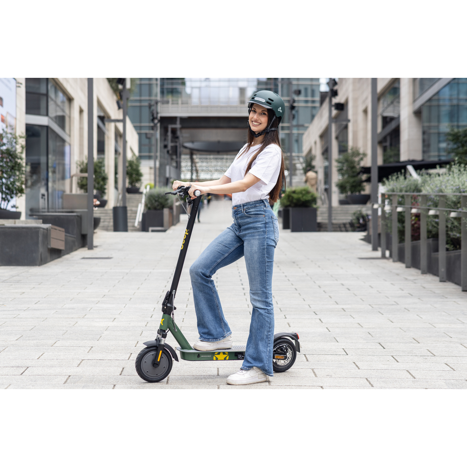 Een vrouw met helm glimlacht terwijl ze op een groene scooter rijdt op een geplaveid pad. Gebouwen op de achtergrond.