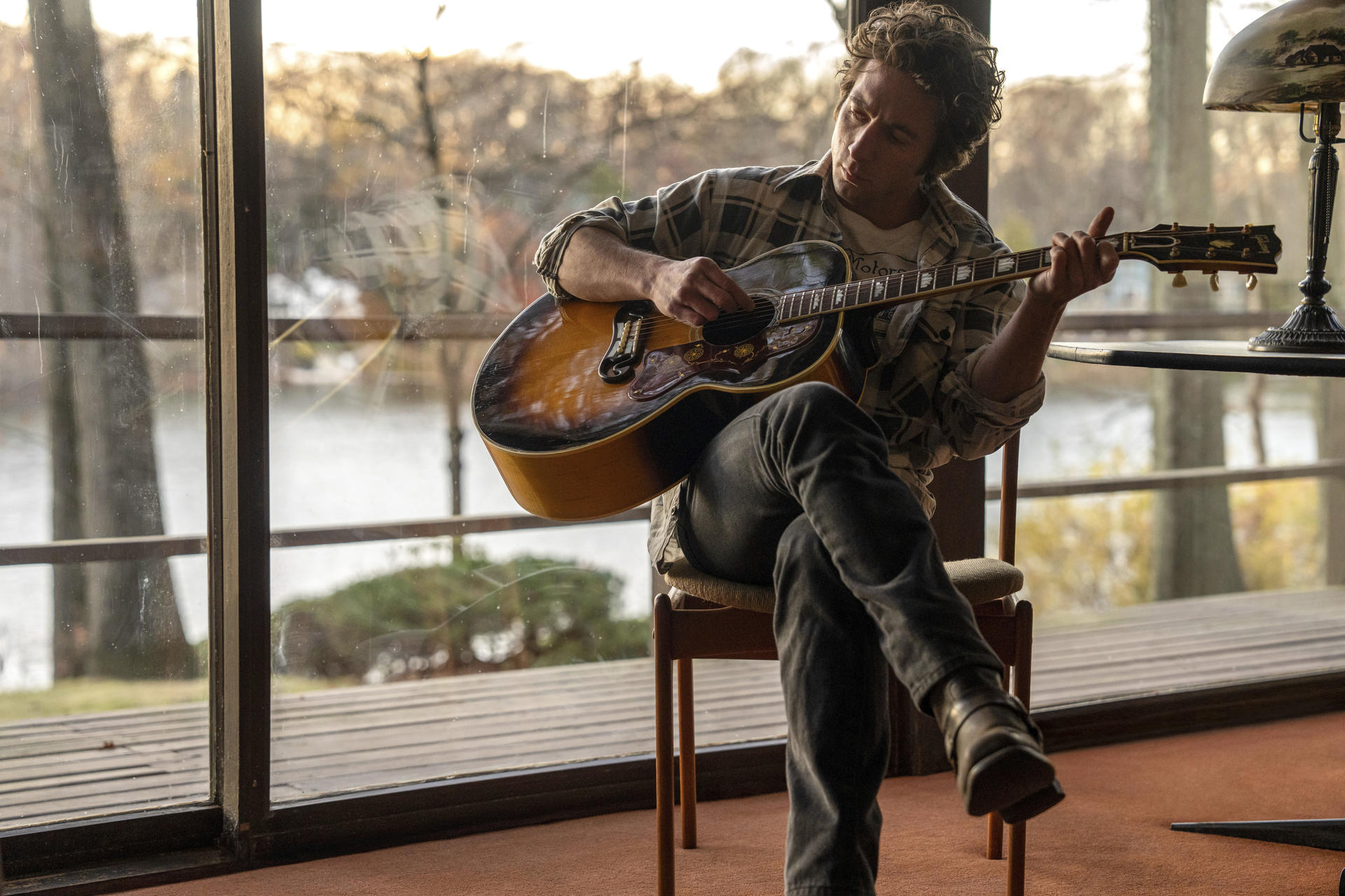 Ein Mann spielt drinnen Gitarre. Er sitzt in der Nähe eines Fensters mit Seeblick, trägt ein kariertes Hemd und Jeans. Eine Lampe steht in der Nähe.