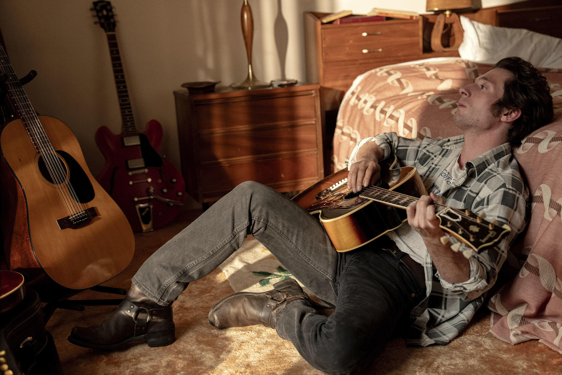 Ein Mann spielt Gitarre in einem Raum. Zwei weitere Gitarren sind in der Nähe. Er trägt ein kariertes Hemd, Jeans und Stiefel und lehnt sich an ein Bett.