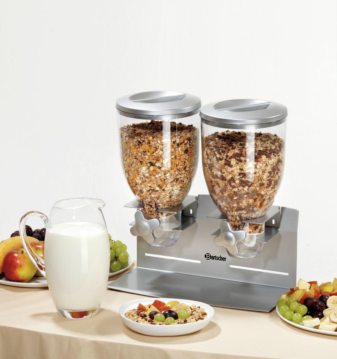 Getreidespender mit Müsli, Obstplatten und einem Milchkanne auf einem Tisch. Weißer Hintergrund.