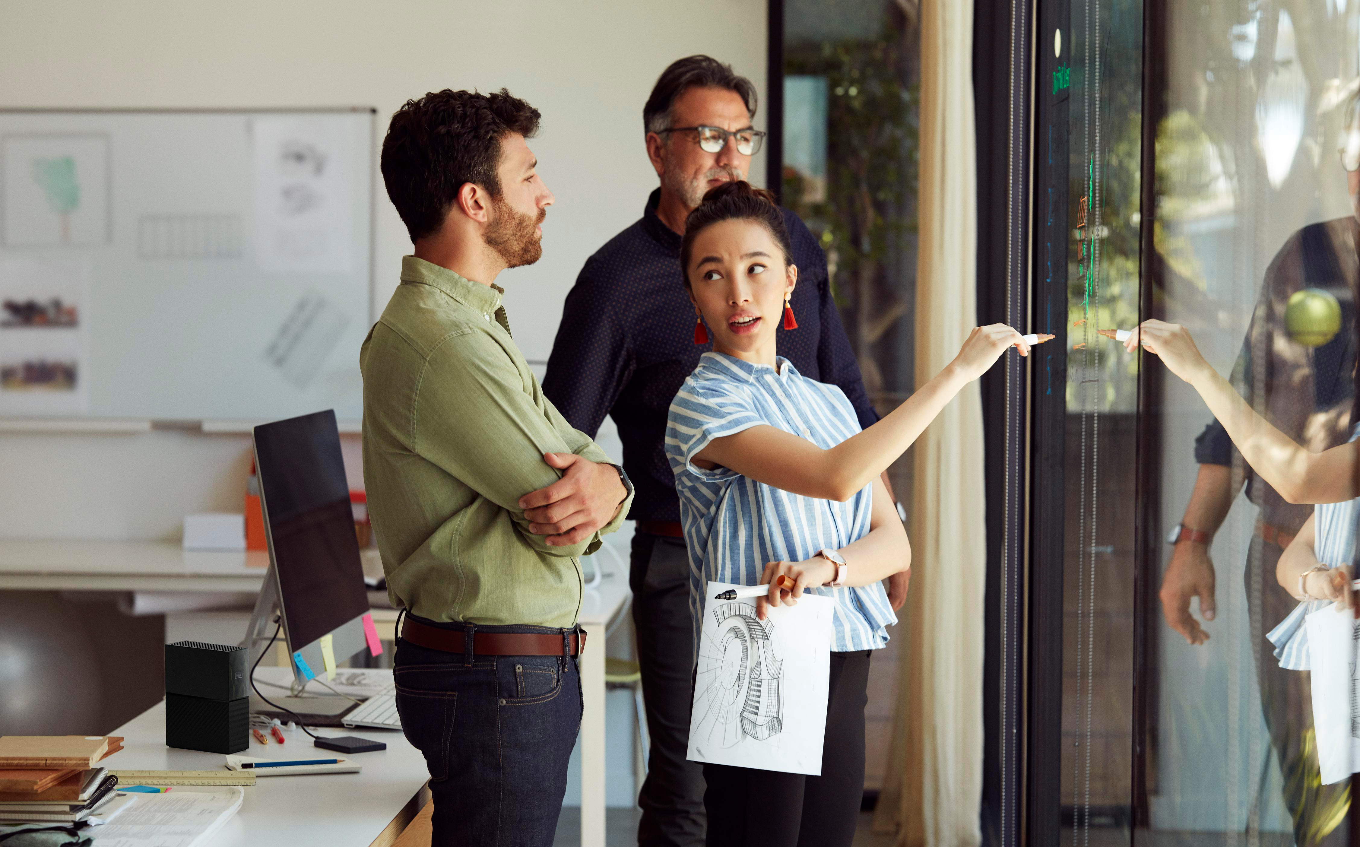 Drei Personen in einem Büro. Eine Frau zeigt auf eine Glaswand, während andere zusehen. Ein Skizze ist in der Hand.