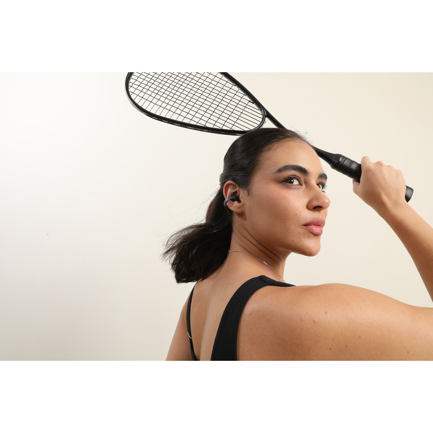 Woman wearing earbuds holds a tennis racket. She wears a black top, looking over her shoulder. Neutral background.