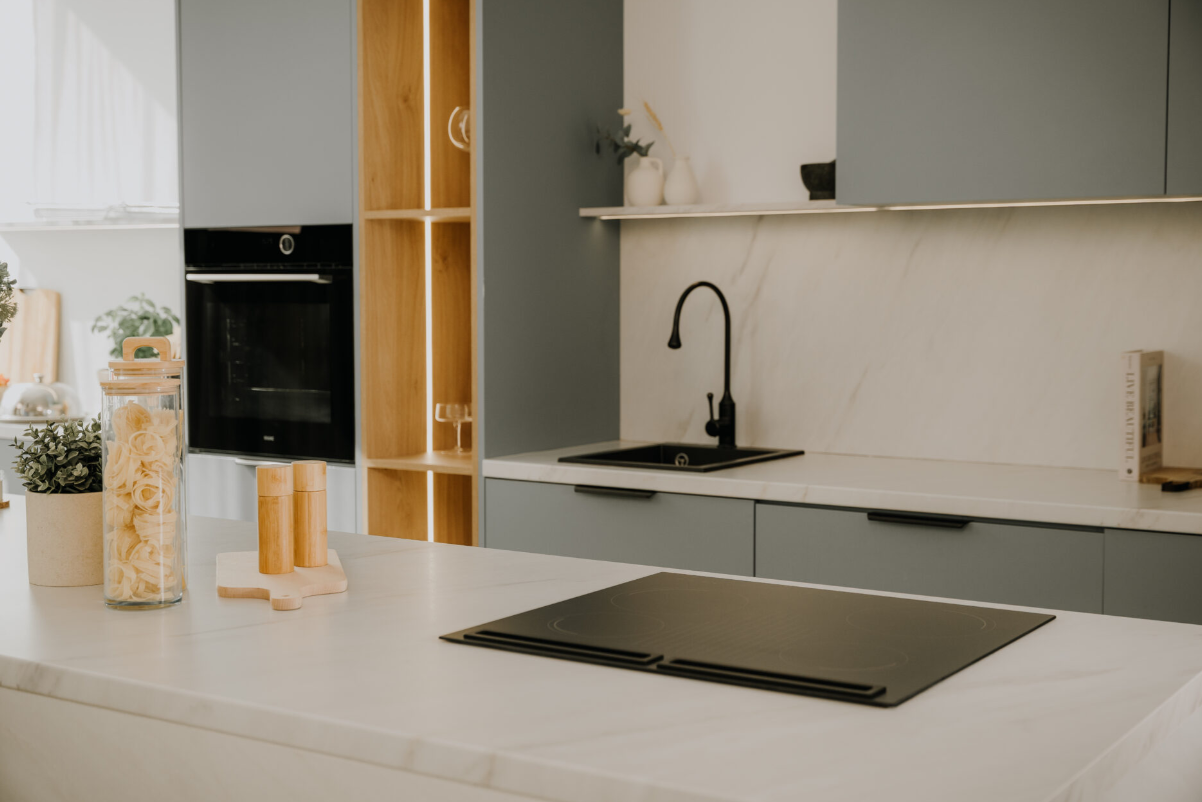 Nowoczesna kuchnia z czarną płytą grzewczą, zlewem i kranem. Zawiera drewniane półki, słoiki i szare szafki. Jasny, czysty design.