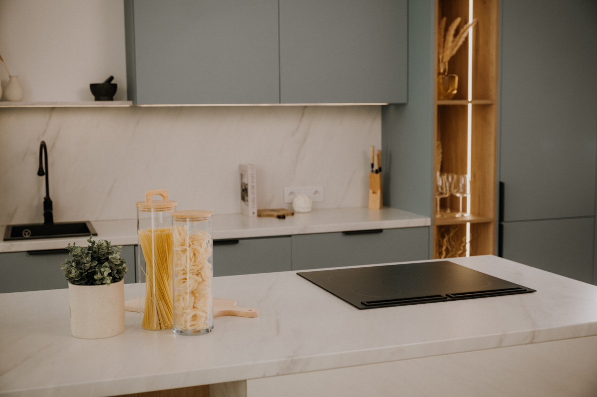 Nowoczesna kuchnia z blatem. Są słoiki makaronu, roślina i płyta grzewcza. Szafki i półki w tle.