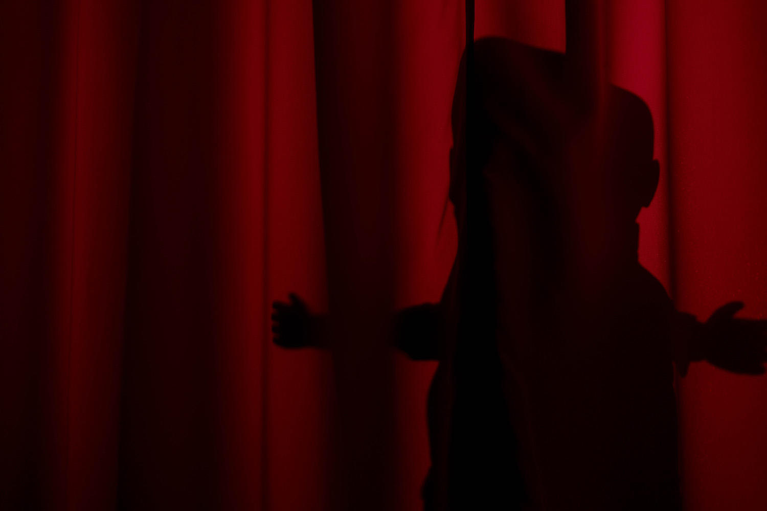 Silhouette einer Person, die einen roten Vorhang öffnet. Die Arme der Person sind ausgestreckt. Der Hintergrund ist dunkelrot.