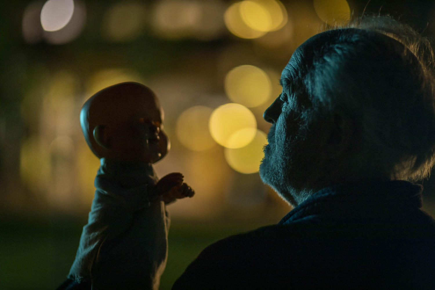 Ein Mann betrachtet eine Puppe. Die Puppe wird hochgehalten. Der Hintergrund hat unscharfe gelbe Lichter.