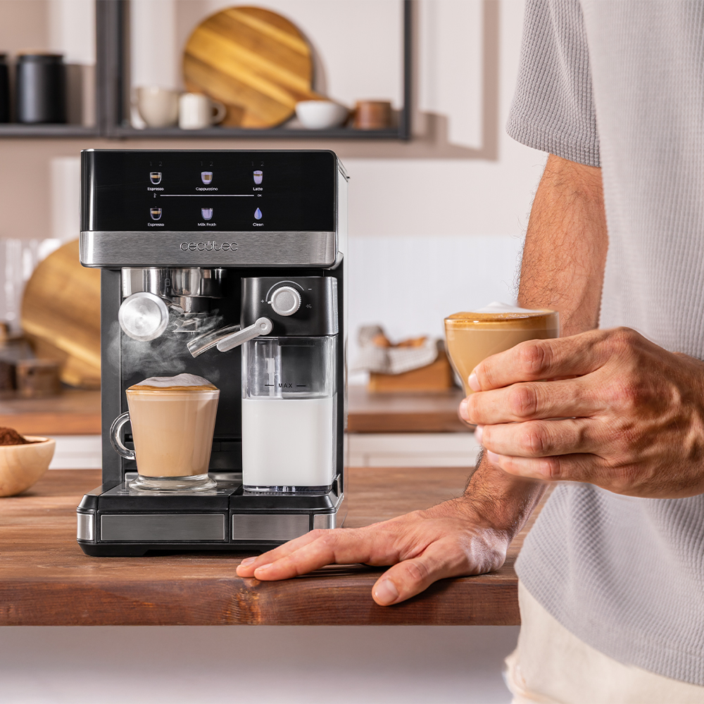 Una persona sostiene un café con espuma cerca de una máquina de café. La máquina tiene un vaso de leche y una taza de café.