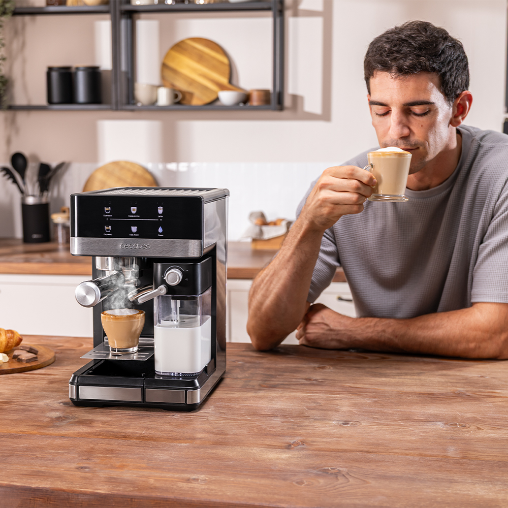 Un hombre bebe café de una taza cerca de una máquina de espresso. Un cruasán y una tabla de madera están en la mesa.