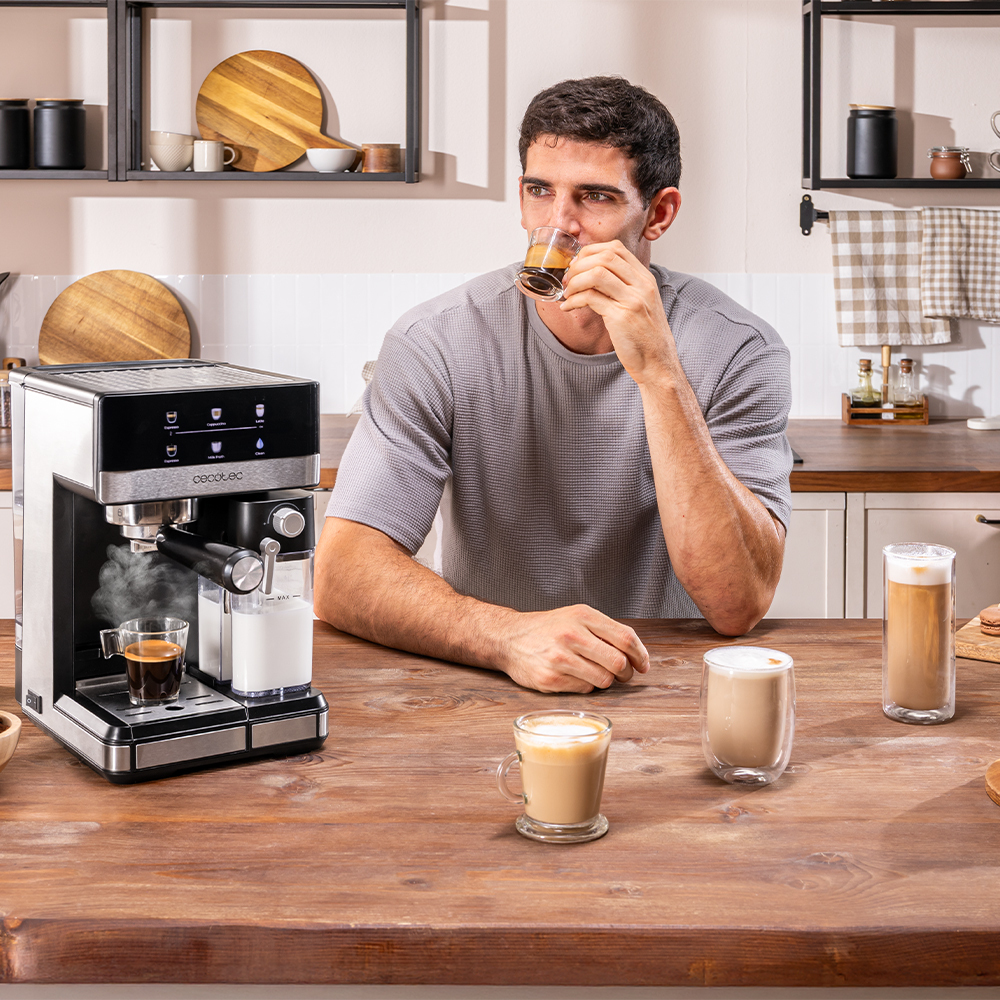 Un hombre bebe café de un vaso mientras está sentado en una mesa de madera con una cafetera y tazas de café. Fondo de cocina.
