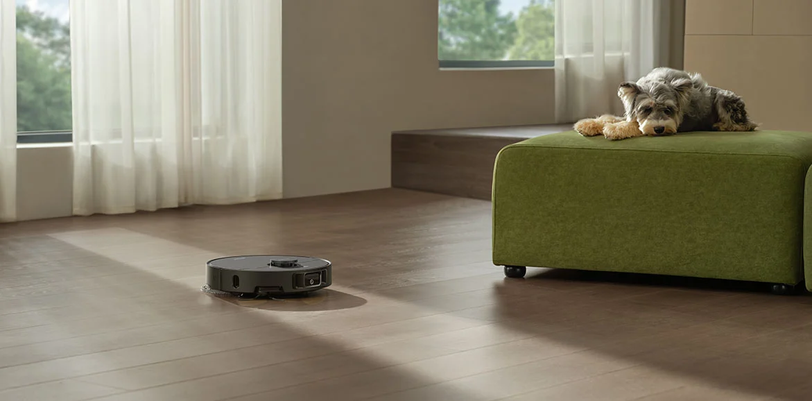 Robot sprząta drewnianą podłogę. Pies odpoczywa na zielonej pufie. Okna z białymi zasłonami po lewej.