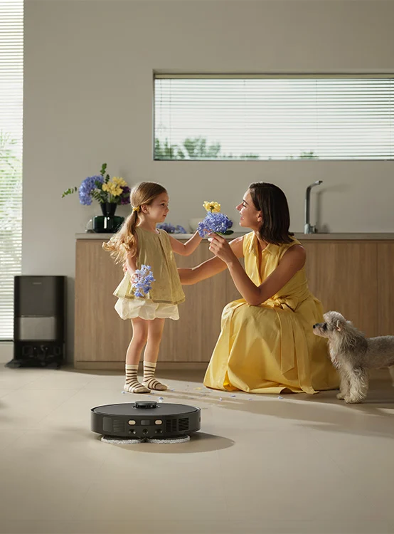 Kobieta i dziewczynka wymieniają się kwiatami w kuchni. Robot sprząta podłogę. Mały pies obserwuje.