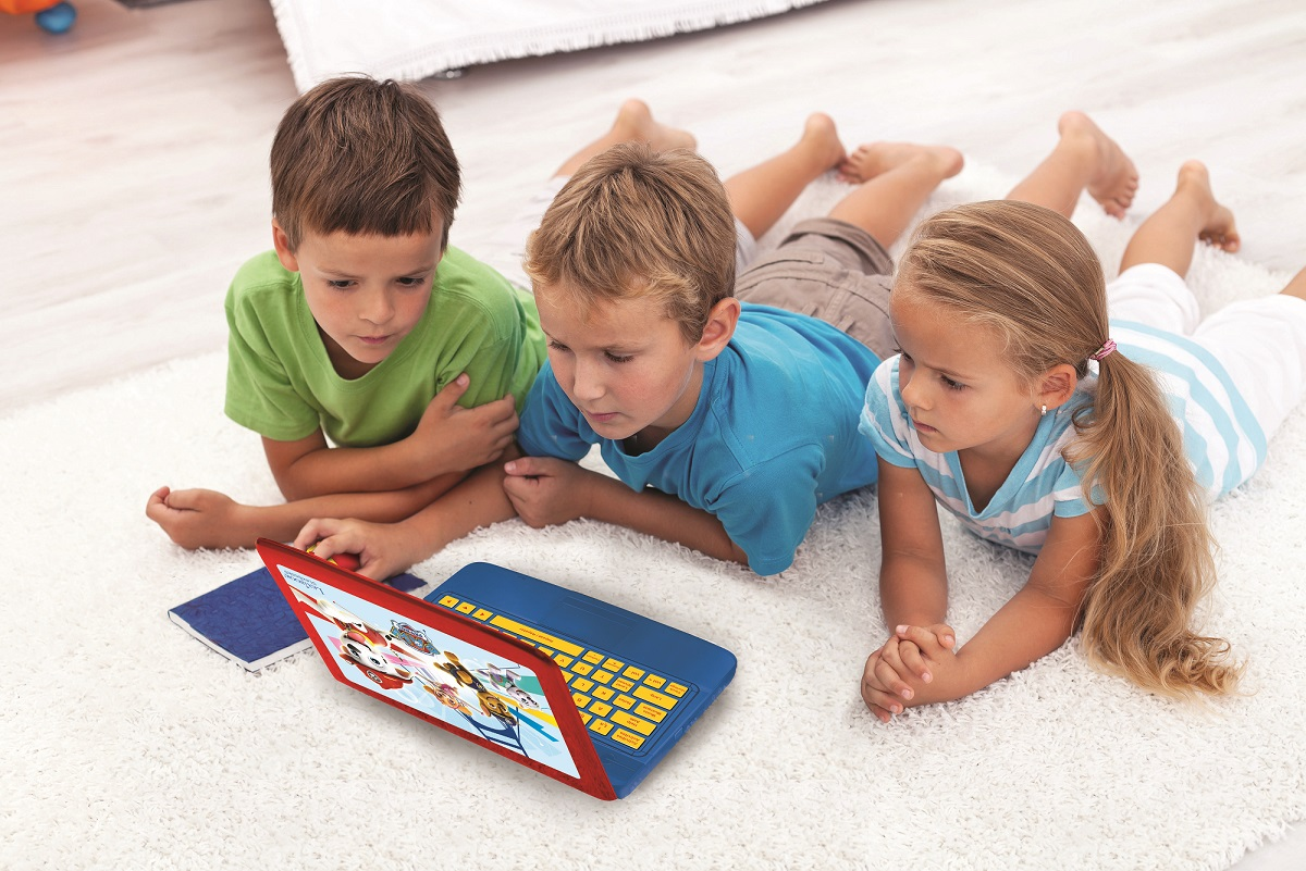 Drei Kinder liegen auf einem weißen Teppich und schauen auf einen blauen Spielzeug-Laptop. Der Laptop zeigt einen Cartoon. Zwei Jungen und ein Mädchen sind anwesend.