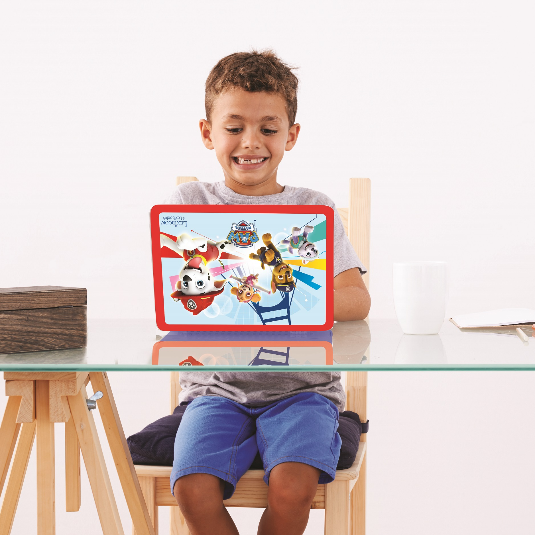 Ein Junge lächelt und betrachtet ein Tablet mit einem Paw Patrol-Design. Er sitzt an einem Glastisch mit einer Holzkiste und einer Tasse.