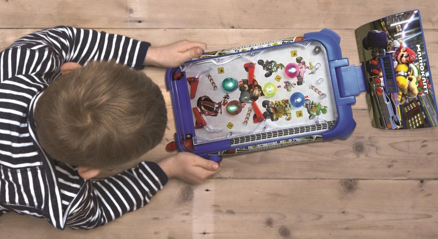 Ein Kind spielt mit einem Mario Kart Flipper. Das Spiel befindet sich auf einer Holzoberfläche. Das Kind trägt ein gestreiftes Hemd. Spielfiguren sind sichtbar.