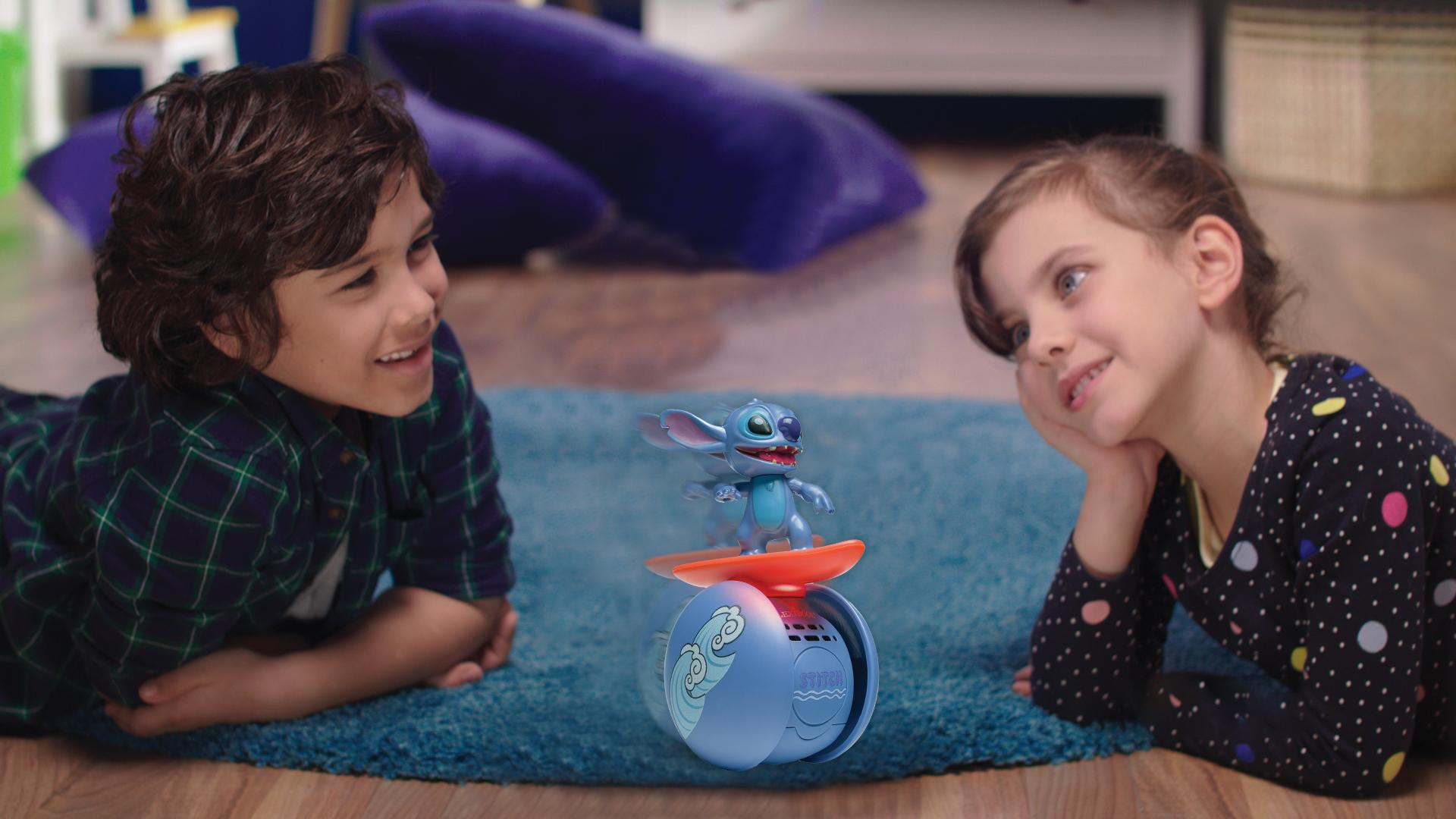 Zwei Kinder betrachten ein Stitch-Spielzeug auf einem blauen Teppich. Das Spielzeug ist auf einem Surfbrett. Im Hintergrund sind Kissen.