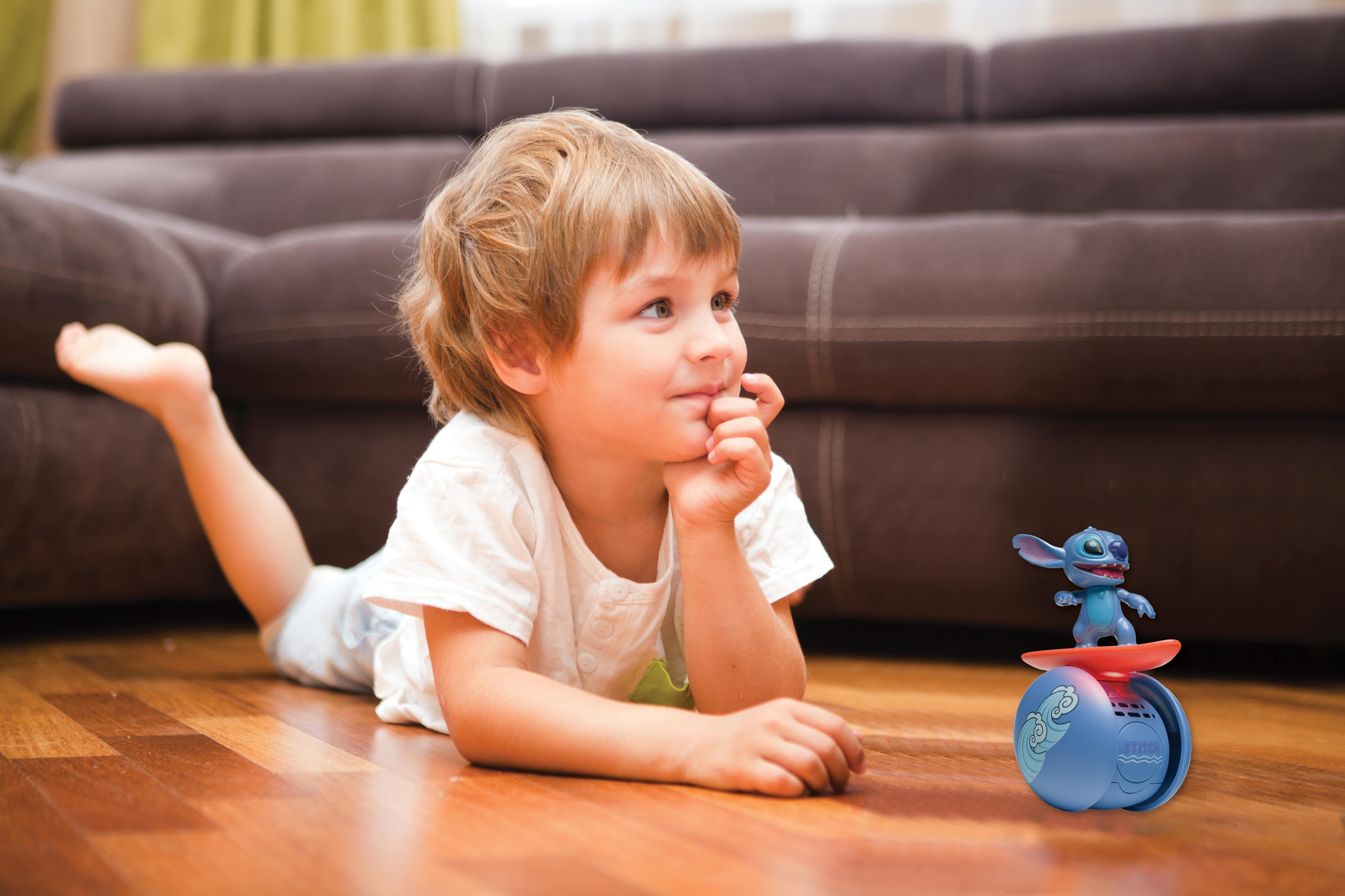 Ein Junge liegt auf einem Holzboden und blickt nach oben. Ein blaues Stitch-Spielzeug steht in der Nähe. Braunes Sofa im Hintergrund.