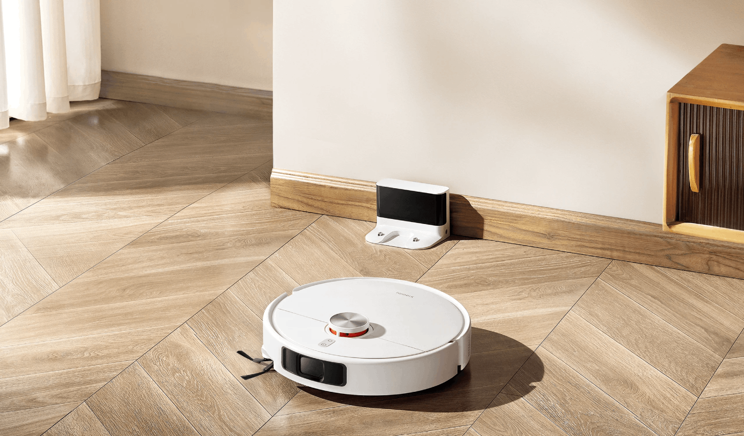Una aspiradora robot blanca está en un piso de madera cerca de su estación de carga. Un armario está a la derecha.