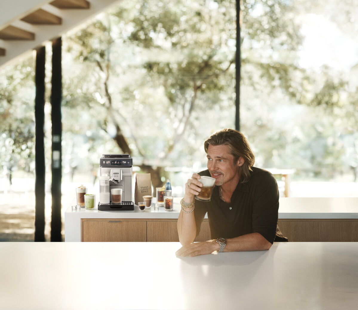 Brad Pitt kávét iszik. Egy kávéfőzőgép, kávéscsészék és kávébabok vannak a pulton. A fák láthatók az ablakon keresztül.