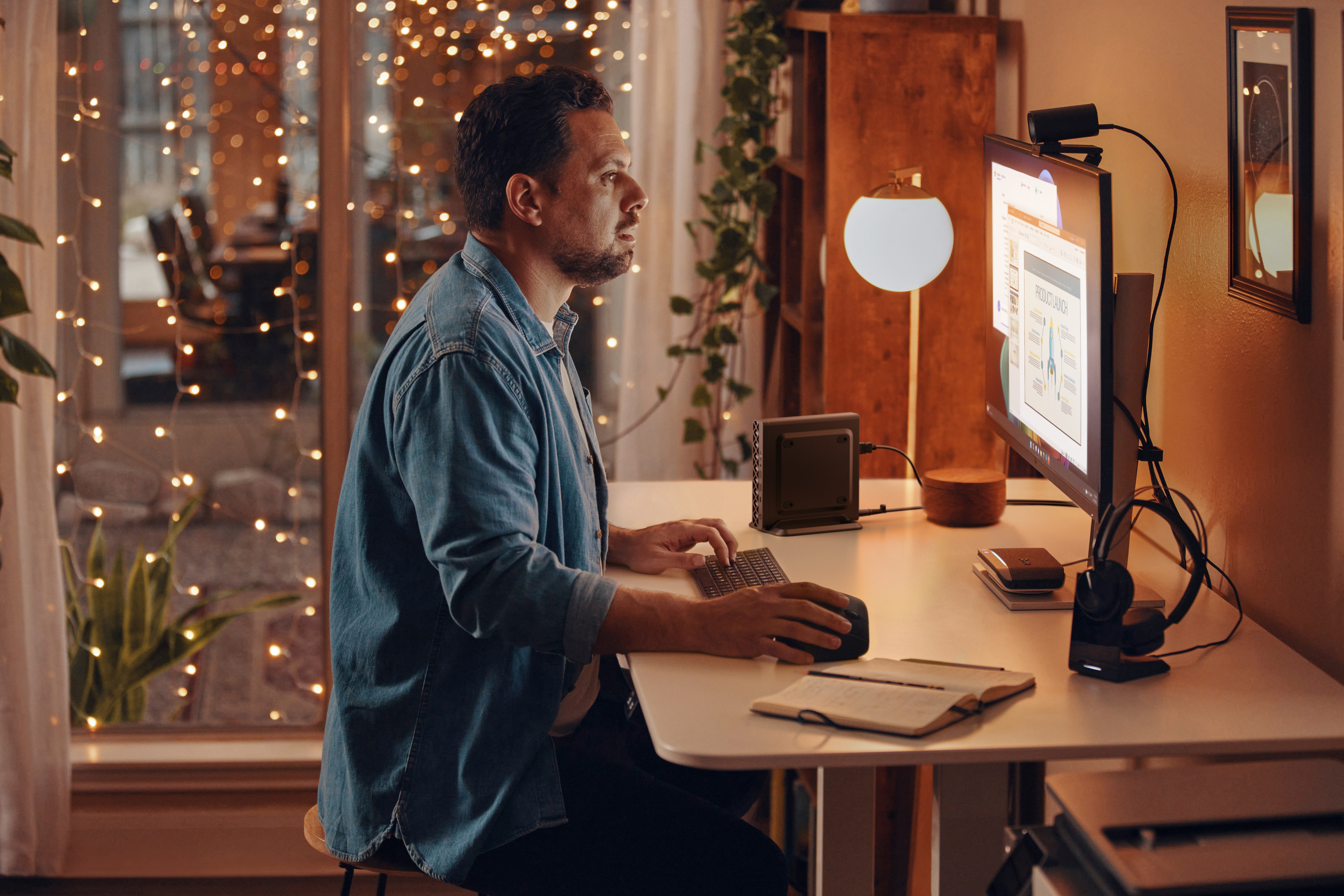 Ein Mann in einem Jeanshemd arbeitet an einem Schreibtisch mit einem Computer. Feenlichter schmücken das Hintergrundfenster. Eine Lampe steht daneben.