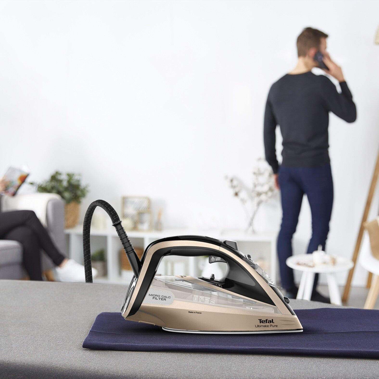 Złote żelazko na niebieskiej tkaninie. Mężczyzna przez telefon, kobieta czyta. Biały pokój, szary stół. Marka Tefal.