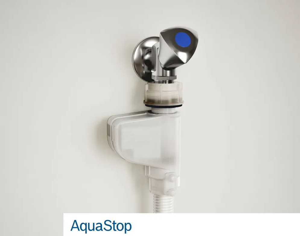 Chromowane urządzenie AquaStop jest zamontowane na białej ścianie. Urządzenie posiada niebieski przycisk i przezroczystą plastikową część.