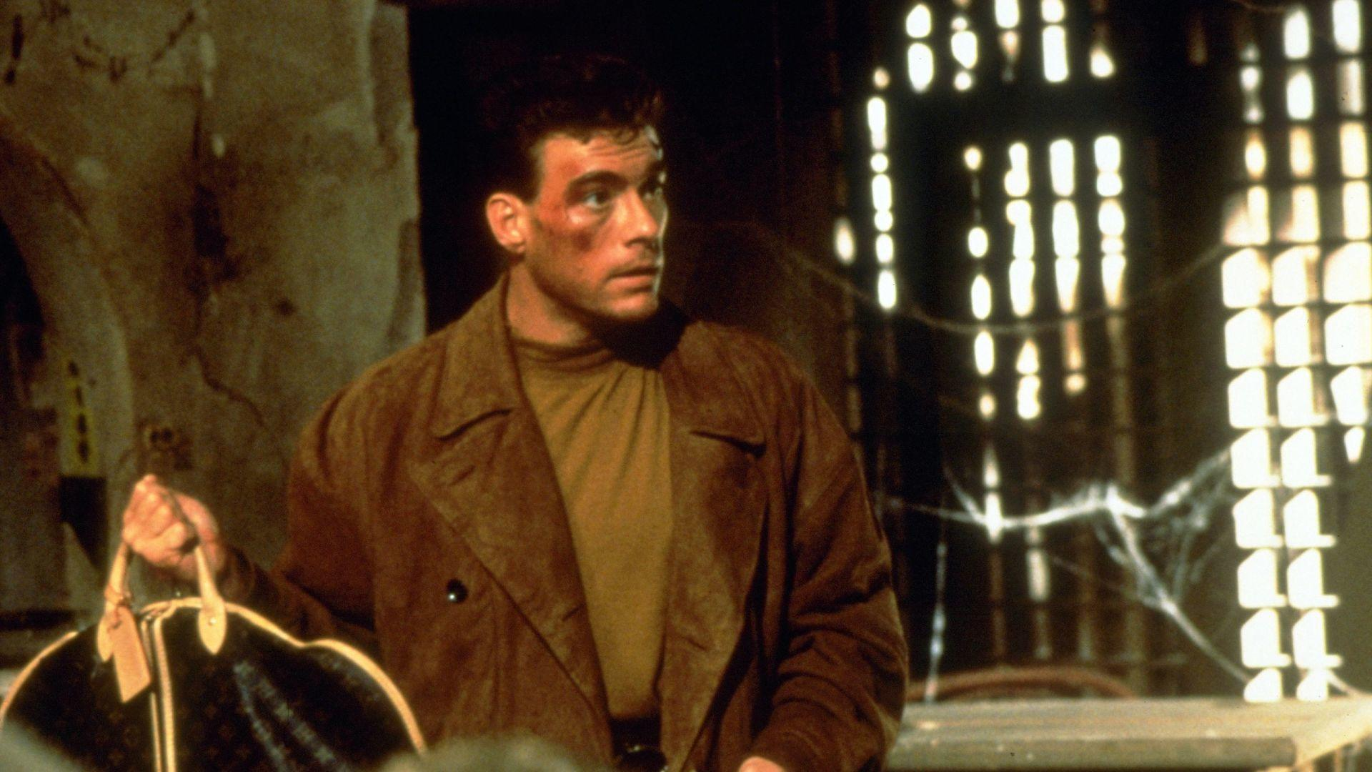 Jean-Claude Van Damme mit einem blauen Fleck hält eine Tasche. Er trägt einen braunen Mantel und ein beiges Hemd. Im Hintergrund ist ein helles Fenster.
