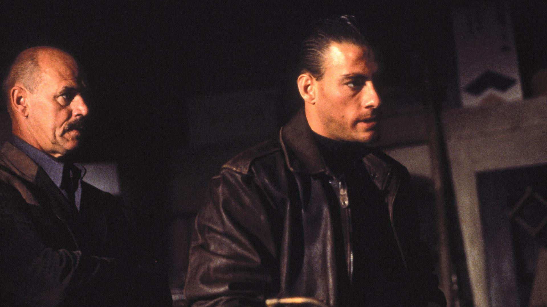 Zwei Männer stehen in einem dunklen Raum. Einer trägt eine Lederjacke, der andere einen Anzug. Beide schauen ernst weg.