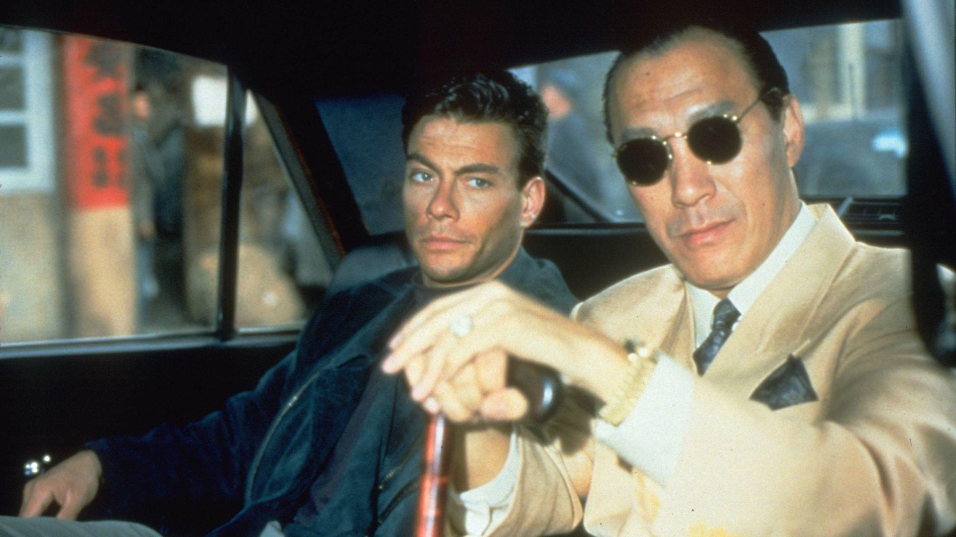Zwei Männer in einem Auto. Einer trägt eine Sonnenbrille und einen Anzug, der andere eine blaue Jacke. Der Hintergrund zeigt eine Straßenszene.