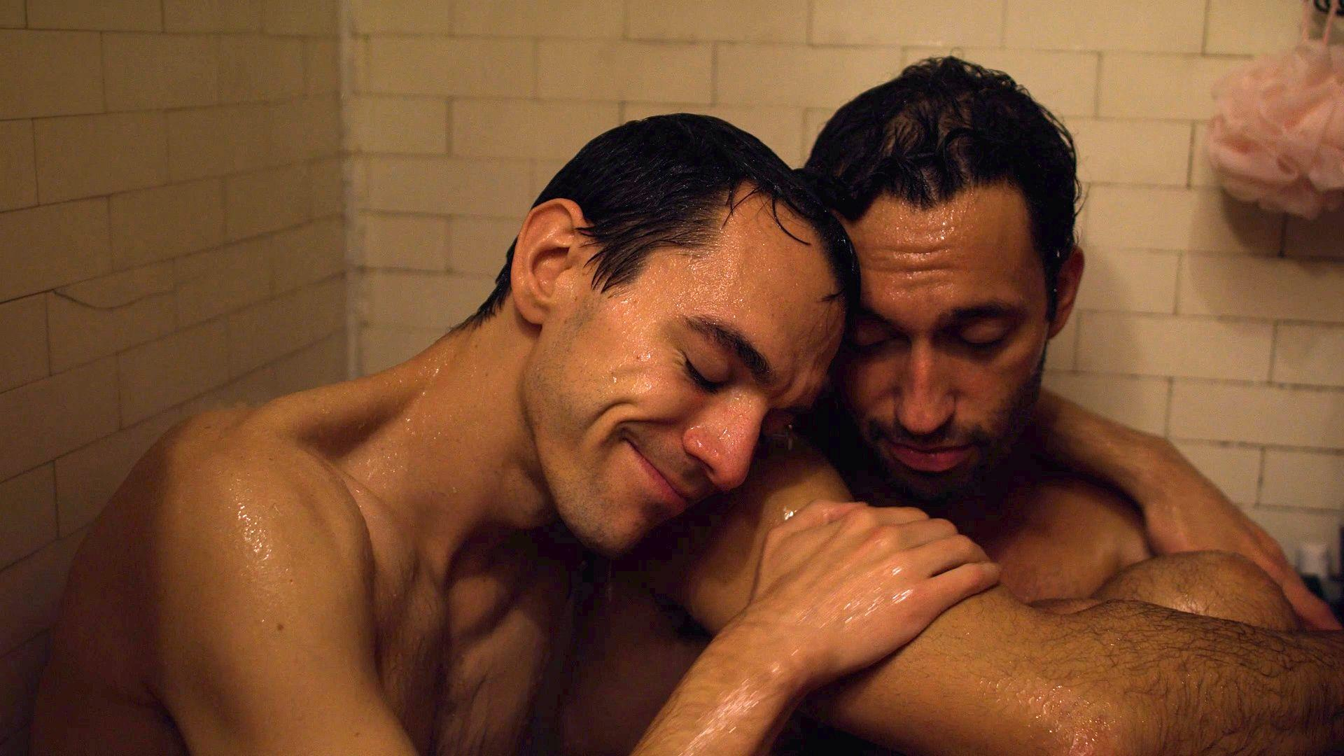 Zwei nasse Männer unter der Dusche. Einer lächelt und lehnt sich an den anderen. Weiße Fliesenwand und ein rosa Schwamm im Hintergrund.