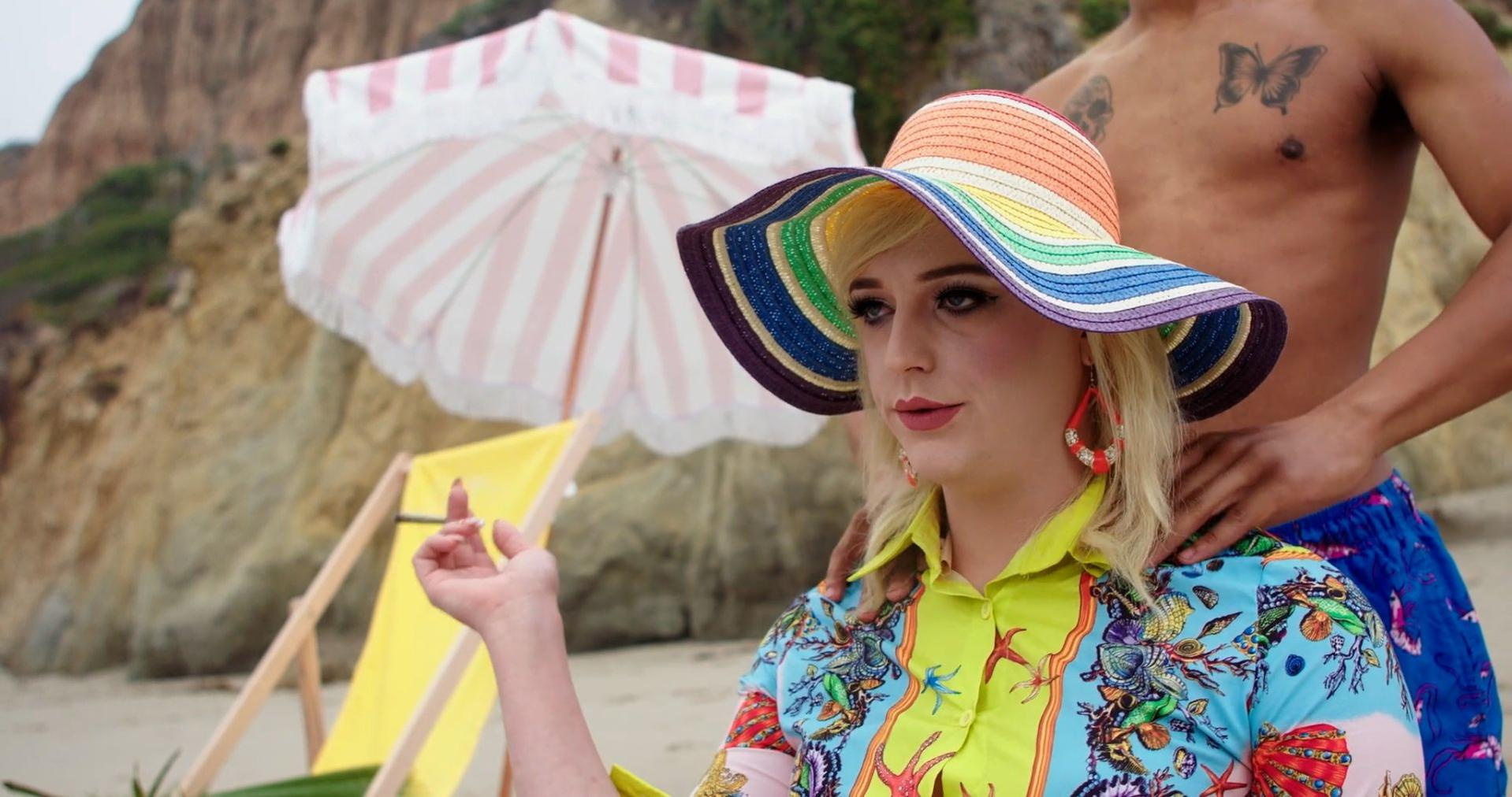 Frau mit Regenbogenhut am Strand, hält Zigarette. Mann mit Schmetterling-Tattoo steht dahinter. Sonnenschirm und Stühle.