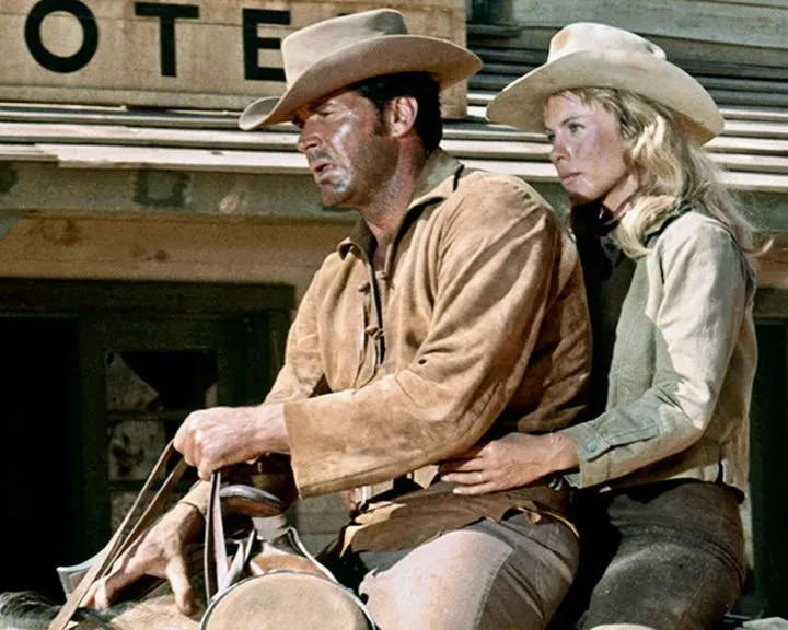 Ein Mann und eine Frau mit Cowboyhüten, der Mann hält Zügel, in der Nähe eines 'OTE'-Schilds. Die Frau legt eine Hand auf seinen Arm.