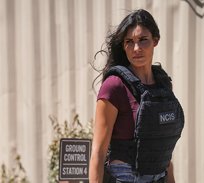 Eine Frau in einer Weste mit der Aufschrift "NCIS" blickt zur Seite. Hinter ihr eine beige Wand und ein Schild mit der Aufschrift "GROUND CONTROL STATION 4".
