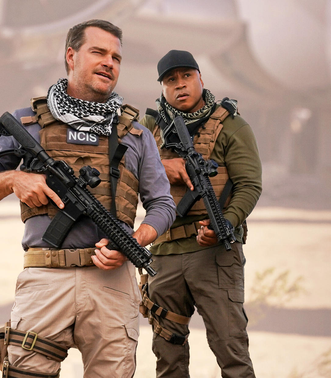 Zwei Männer in taktischer Ausrüstung halten Gewehre. Einer trägt Schal und Weste mit 'NCIS'. Der andere trägt eine schwarze Kappe.