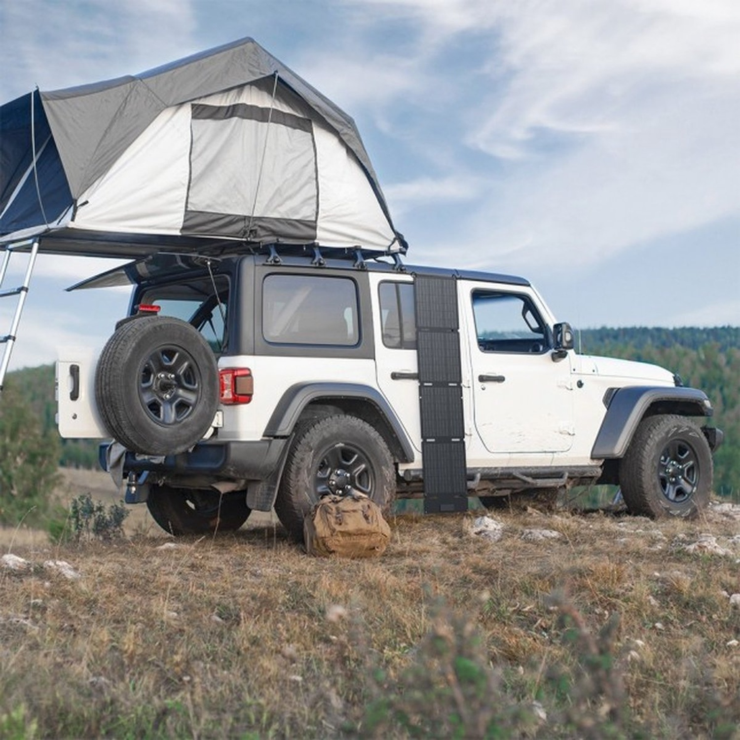 A white Jeep with a rooftop tent is parked on a hill. A spare tire is mounted on the back, with a backpack nearby.
