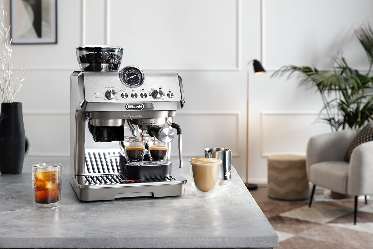 Espresszó gép kávéval, jeges kávé pohárral és tejeskávéval. A háttérben lámpa, szék és növény látható.