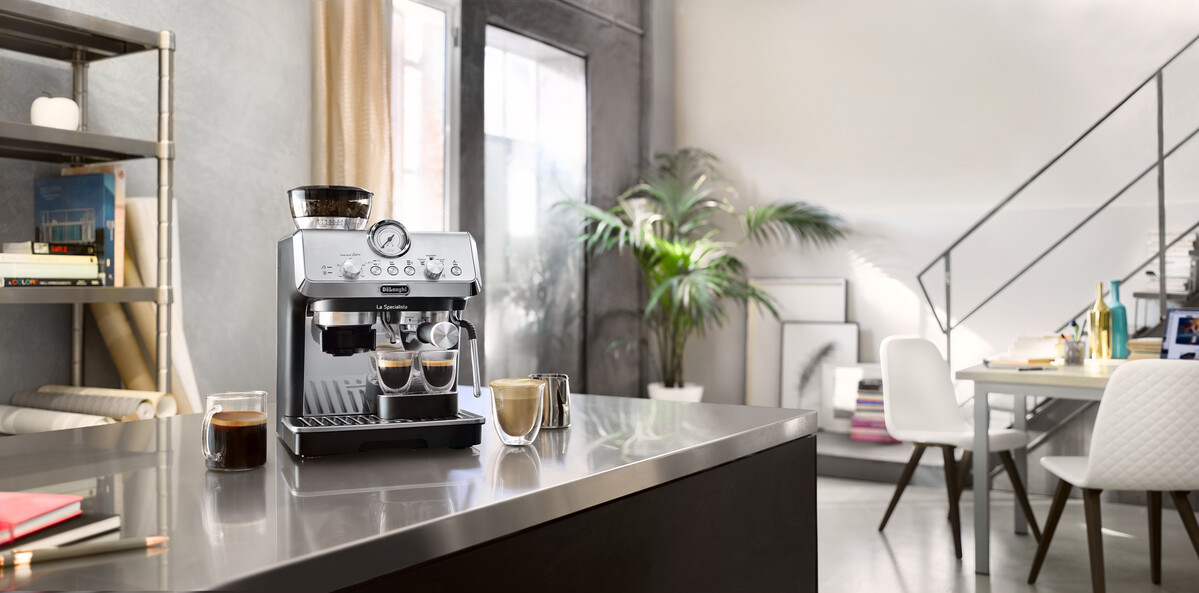 Espresszógép a pulton, kávéscsészékkel. Egy növény és egy asztal székekkel a háttérben. A szoba jól megvilágított.